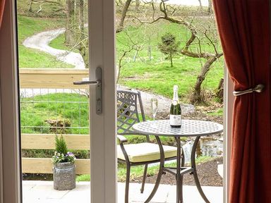 A metal table and chair set with a bottle and glass on the table outside a door with green grass and trees beyond at Park Brook Retreat in Scorton