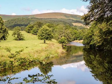 A river flowing beside a grassy field and trees with hills in the background at Park Brook Retreat in Scorton