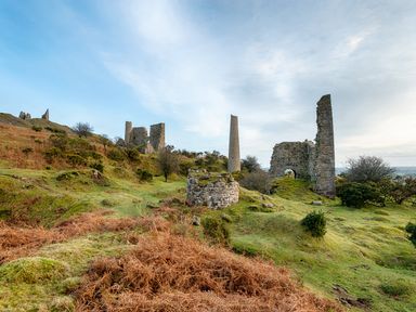 Ruins of stone buildings and towers on grassy hills with shrubs under a blue sky