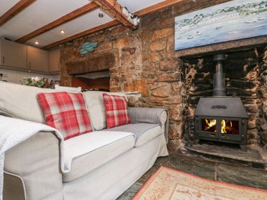 A living room with a beige sofa and red plaid cushions near a wood burning stove in a stone fireplace at Tinners Cottage in Liskeard
