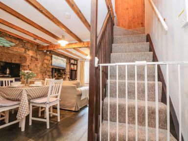 A carpeted staircase with a safety gate next to a dining area with a table and chairs in a room with exposed wooden beams and stone walls at Tinners Cottage in Liskeard