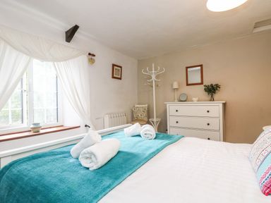 A bedroom with a bed covered by a white sheet and teal blanket with rolled towels on top next to a window with white curtains a white dresser with a lamp and a mirror and a coat rack at Tinners Cottage in Liskeard