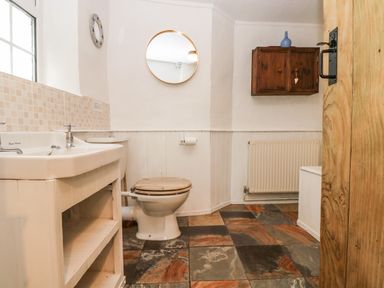 A bathroom with a sink a toilet with wooden seat a wall cabinet and a round mirror at Tinners Cottage in Liskeard