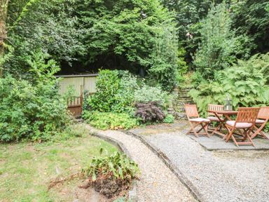 A garden area with a gravel path wooden outdoor table and chairs surrounded by plants at Tinners Cottage in Liskeard