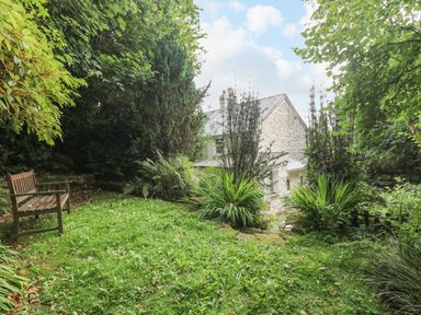 A garden area with grass a wooden bench various plants and trees and a stone house in the background at Tinners Cottage in Liskeard