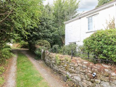 A stone wall with black metal railing next to a gravel driveway and a white building partially visible behind trees and bushes at Tinners Cottage in Liskeard