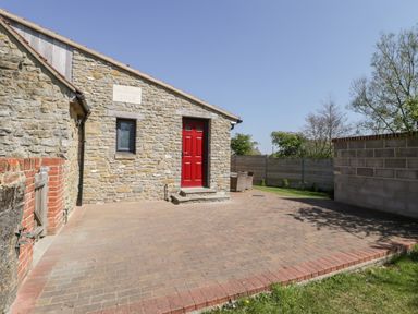 A stone house with a red door and small window on a paved patio area with outdoor chairs and a wooden fence at The Stone Barn in Evercreech near Shepton Mallet