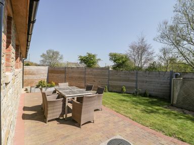 An outdoor patio area with a wooden table and six wicker chairs beside a lawn and wooden fence at The Stone Barn in Evercreech near Shepton Mallet