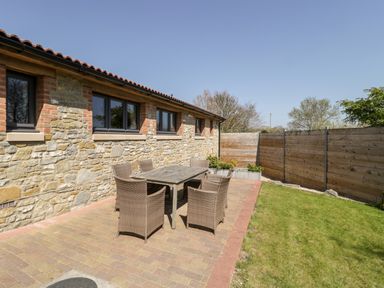 An outdoor patio with a table and six chairs next to a stone building with windows and a wooden fence along the grass at The Stone Barn in Evercreech near Shepton Mallet