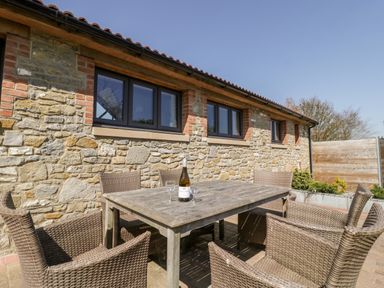 An outdoor patio area with a wooden table and wicker chairs against a stone wall at The Stone Barn in Evercreech near Shepton Mallet