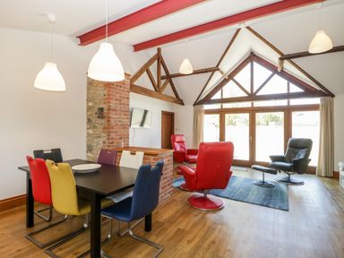 A living room with multicolored dining chairs a black table red and black armchairs a blue rug wooden beams and large windows at The Stone Barn in Evercreech near Shepton Mallet