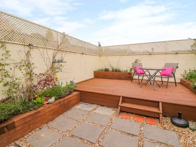 An outdoor patio with wooden decking, two chairs with pink cushions, a small table, and planter boxes with plants at Bank House Capel Iwan near Newcastle Emlyn