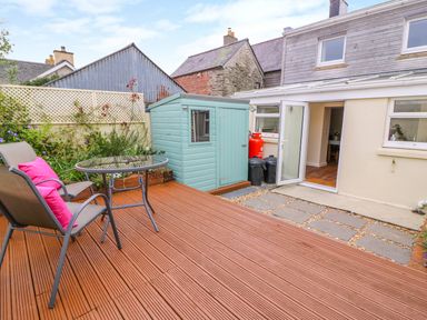 A small patio with wooden decking a round glass table two chairs with pink cushions a garden shed and open glass door at Bank House in Capel Iwan near Newcastle Emlyn