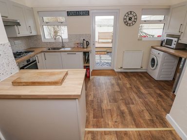 A kitchen with wooden countertops a chopping board a sink a stove a kettle a washing machine a microwave a wall clock and a door leading outside at Bank House in Capel Iwan near Newcastle Emlyn