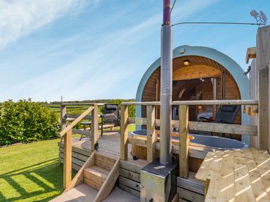 An outdoor wooden deck with chairs a hot tub and a round wooden cabin in a grassy area at Two Hoots in Northop