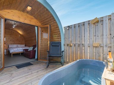 A wooden pod bedroom open to a deck with a hot tub and chair at Two Hoots in Northop
