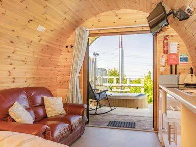 A wooden cabin interior with a red leather sofa, pillows, kitchenette, door open to a deck with a chair and a small hot tub at Two Hoots in Northop
