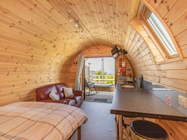 A wooden pod interior with a bed a red sofa a kitchen counter stools and an open door leading to a small deck with a hot tub and chair at Two Hoots in Northop
