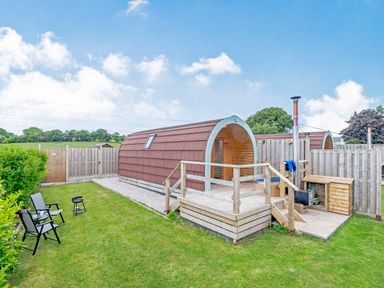 A wooden pod with a deck and chimney in a fenced grassy garden with chairs and a small grill at Ewe Two in Oakenholt near Northop