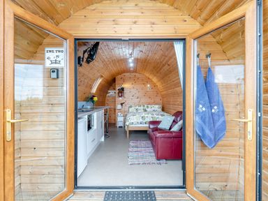 Interior of a wooden pod with double doors open showing a kitchenette a bed a red sofa and two towels hanging at Ewe Two in Oakenholt near Northop