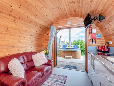 An interior wooden cabin with a red leather sofa, kitchen area, wall-mounted TV, and an outdoor hot tub on a wooden deck at Ewe Two in Oakenholt near Northop