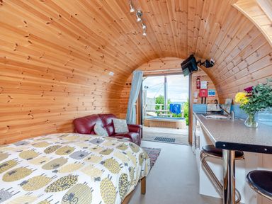 An interior of a wooden pod with a bed with patterned bedding a red leather couch a kitchenette with a countertop and stools and a door leading to an outdoor deck with a hot tub at Ewe Two in Oakenholt near Northop