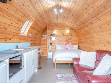 An interior of a wooden cabin with a bed with pillows a red sofa with cushions a kitchen counter with a sink and bar stools at Ewe Two in Oakenholt near Northop