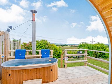 An outdoor wooden hot tub on a deck with towels hanging on a wooden railing and a view of a grassy field at Ewe Two in Oakenholt near Northop
