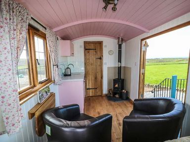 A small room with two armchairs a wood stove a kitchenette and windows with floral curtains at Llety'r Bugail in Holyhead