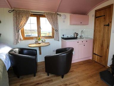 A small kitchen area with pink cabinets and a kettle next to two black armchairs and a wooden table under a window at Llety'r Bugail in Holyhead