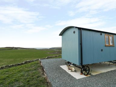 A blue shepherd's hut on wheels on a gravel pad with a grassy field and hills in the background at Llety'r Bugail in Holyhead