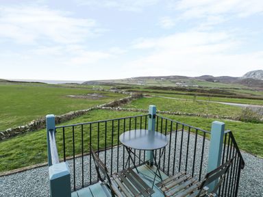 A small balcony with a round table and two chairs overlooking green fields and hills at Llety'r Bugail in Holyhead