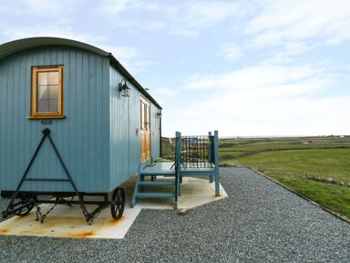 A blue wooden caravan with stairs and railings on a gravel area with green fields in the background at Llety'r Bugail in Holyhead