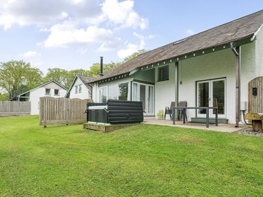 An outdoor patio with chairs and a hot tub in front of a white house with a grassy yard at Elm - Woodland Cottages in Bowness-on-Windermere