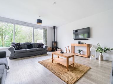 A living room with gray sofas wooden coffee table tv on wall and large windows at Elm - Woodland Cottages in Bowness-on-Windermere