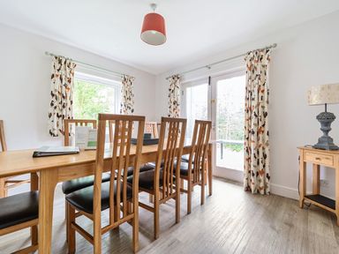 A dining room with a wooden table and six chairs near windows with patterned curtains at Elm - Woodland Cottages in Bowness-on-Windermere
