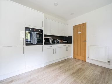 A kitchen with white cabinets a built in oven a microwave kettle toaster wooden door and radiator at Elm - Woodland Cottages in Bowness-on-Windermere
