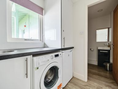 A laundry room with a washing machine dishwasher countertop window and a view into a bathroom at Elm - Woodland Cottages in Bowness-on-Windermere
