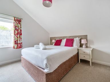 A bedroom with a bed with white linens and red pillows two bedside tables with lamps and a window with floral curtains at Elm - Woodland Cottages in Bowness-on-Windermere