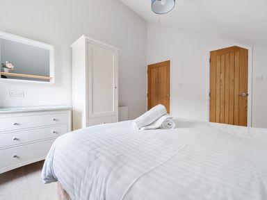 A bedroom with a bed covered in white linens two wooden doors a white wardrobe and white chest of drawers with a mirror at Elm - Woodland Cottages in Bowness-on-Windermere