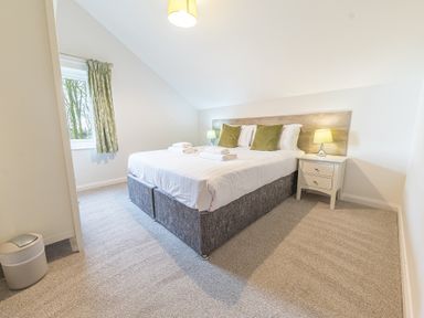 A bedroom with a double bed with white bedding and two green pillows a nightstand with a lamp and a window with green curtains at Elm - Woodland Cottages in Bowness-on-Windermere