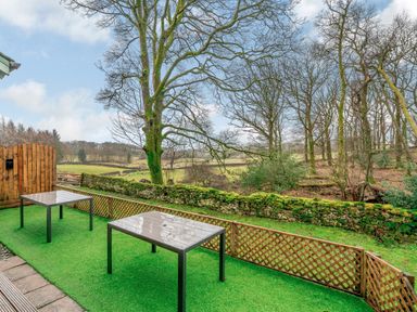 An outdoor patio area with two tables and green artificial grass overlooking a stone wall and trees at Cherry - Woodland Cottages in Bowness-in-Windermere