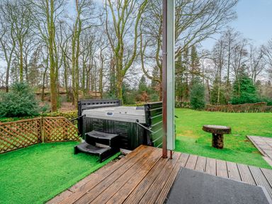 An outdoor area with a hot tub on artificial grass and wooden decking surrounded by trees at Cherry - Woodland Cottages in Bowness-in-Windermere