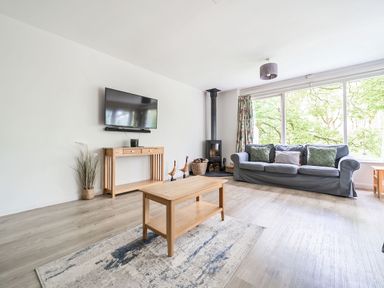 A living room with a gray sofa, wooden coffee table, wall-mounted television, wood-burning stove, and large windows at Cherry - Woodland Cottages in Bowness-in-Windermere