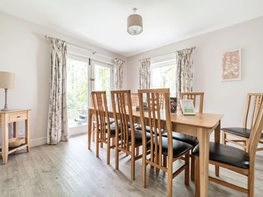 A dining room with a wooden table and eight chairs next to windows with curtains at Cherry - Woodland Cottages in Bowness-in-Windermere