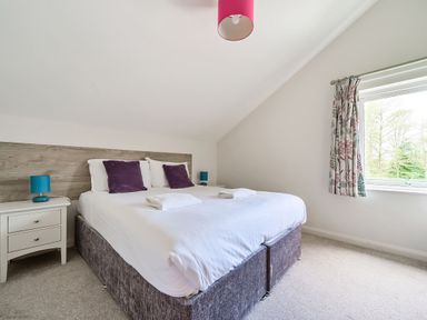 A bedroom with a bed with white linens and purple pillows two bedside tables with blue lamps and a window with patterned curtains at Cherry - Woodland Cottages in Bowness-in-Windermere