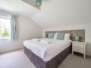 A bedroom with a double bed with white linens and blue pillows next to two white nightstands with lamps and a window with curtains at Cherry - Woodland Cottages in Bowness-in-Windermere