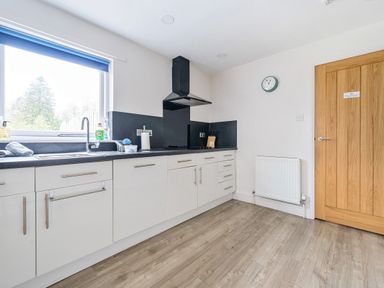 A kitchen with white cabinets a black countertop a window with a blue blind a stove an extractor fan a wooden door and a wall clock at Maple - Woodland Cottages in Bowness-on-Windermere