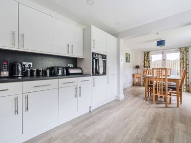 A kitchen with white cabinets and black countertop next to a dining area with wooden table and chairs at Maple - Woodland Cottages in Bowness-on-Windermere