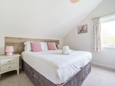 A bedroom with a double bed with pink cushions and white linens two bedside tables with pink lamps a window with patterned curtains and a rabbit painting on the wall at Maple - Woodland Cottages in Bowness-on-Windermere